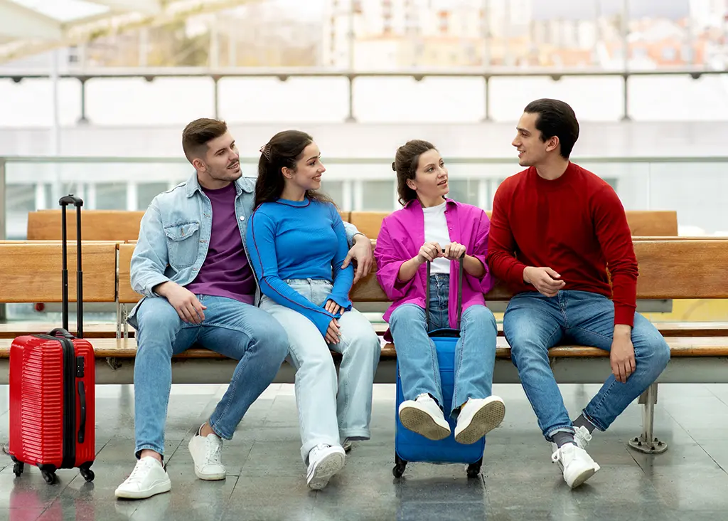 Four friends with red and blue luggage talking on a bench in a bright transit terminal while using sports travel management services.